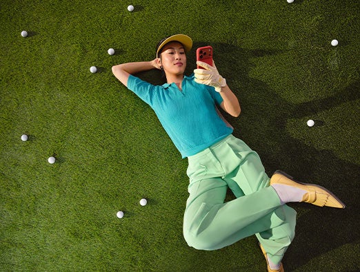 Person lying on their back on a lawn scattered with golf balls, looking at a smartphone.